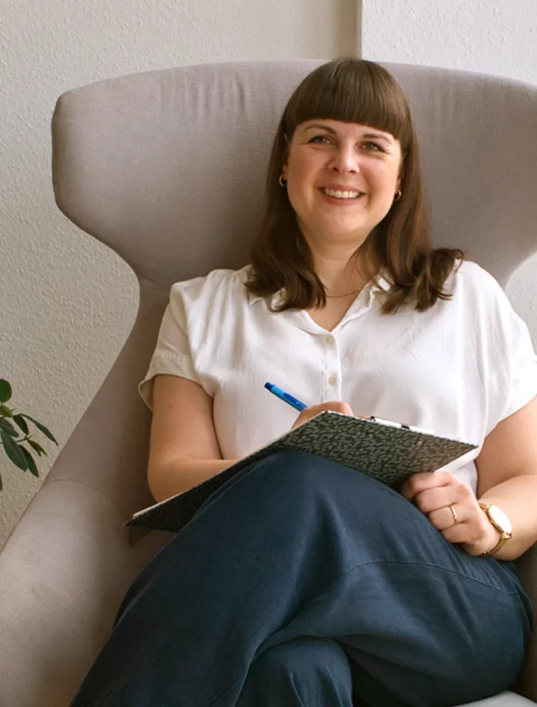 Psychotherapeutin Stella Heimsch sitzt lächelnd mit einem Klemmbrett in einem grauen Sessel.