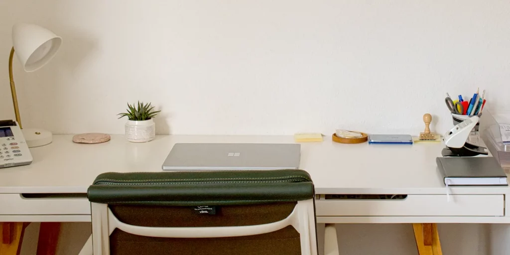 Ordentlicher, weißer Schreibtisch mit Laptop, Lampe und Arbeitsutensilien in einer hellen Praxis.