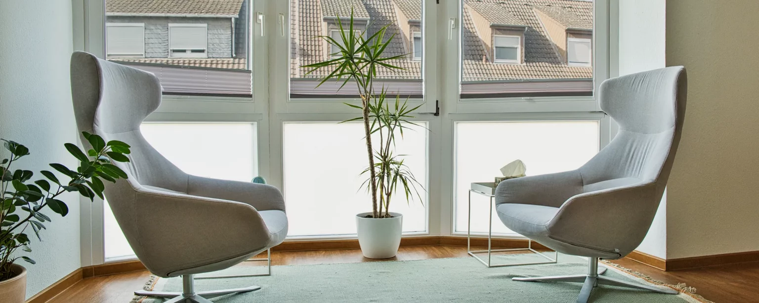 Heller Therapieraum mit zwei modernen grauen Sesseln vor einem großen Fenster mit Blick auf Dächer.