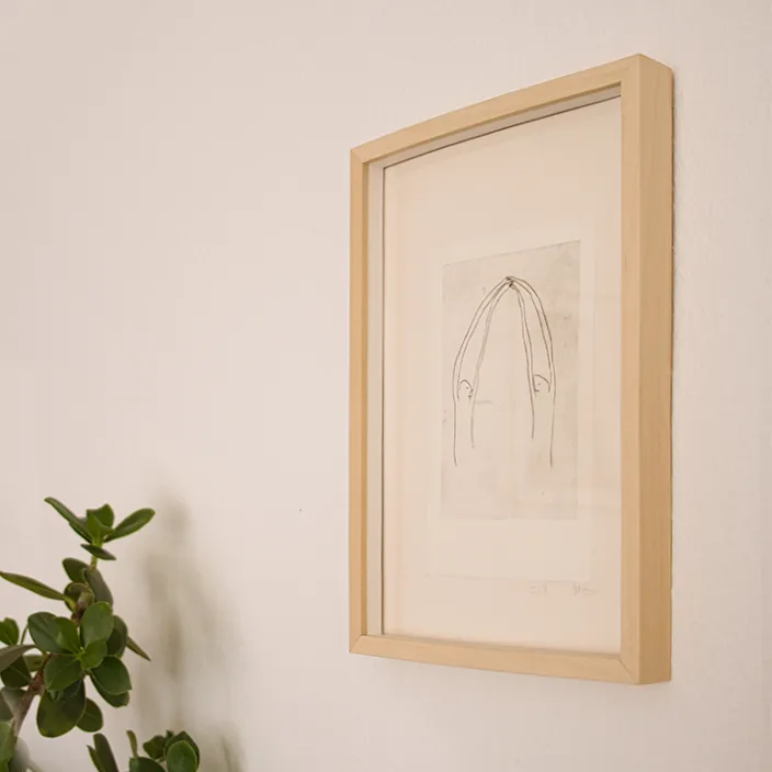 Minimalistische Wanddekoration in einem hellen Holzrahmen neben einer grünen Zimmerpflanze.