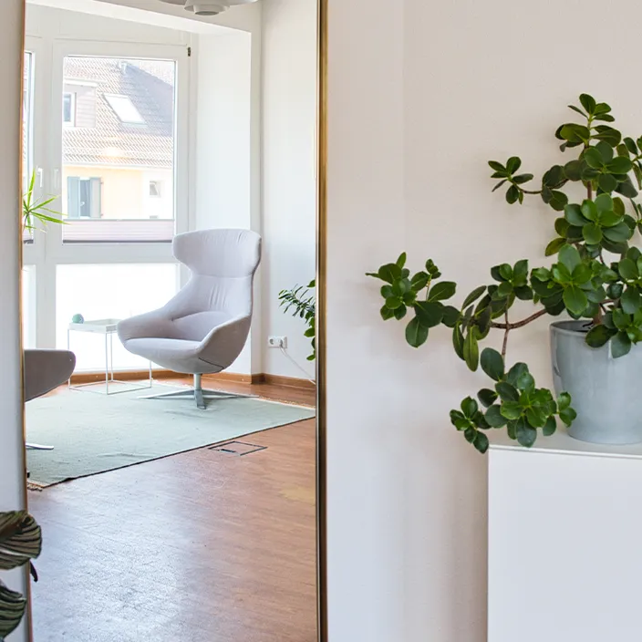 Spiegelreflexion eines hellen Behandlungszimmers mit einem grauen Sessel und einer grünen Pflanze im Vordergrund.